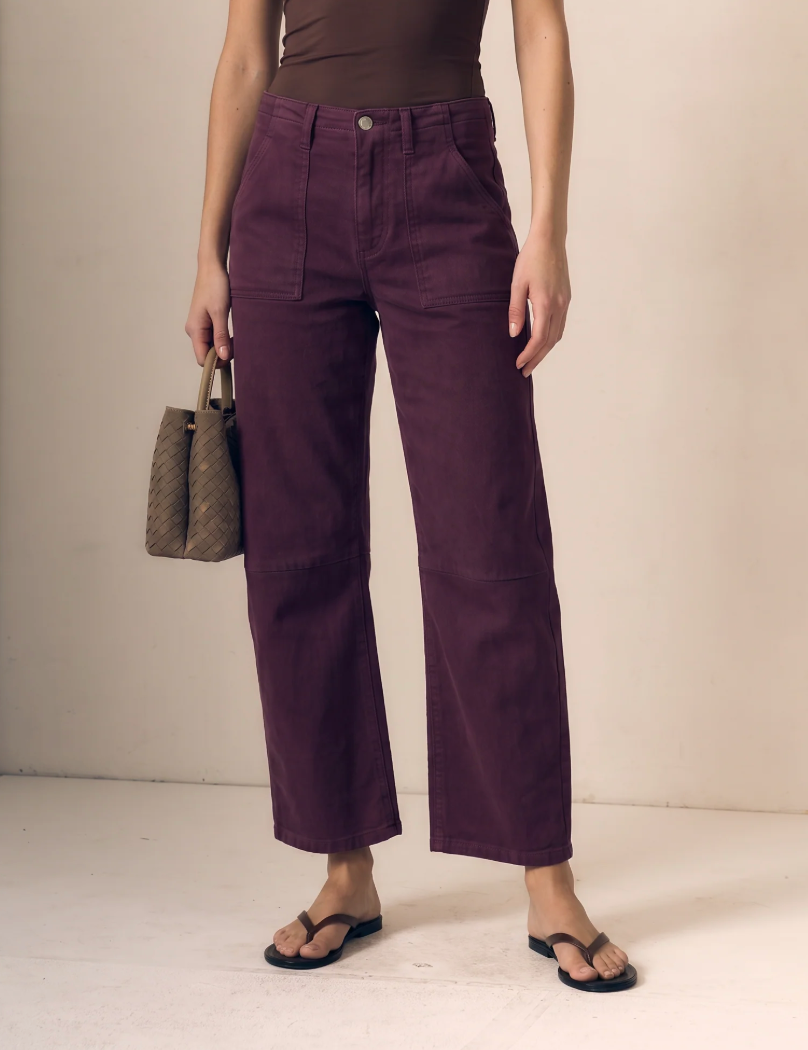 A model wearing a pair of dark purple cropped wide-leg pants, styled with a brown top, black flip-flops, and holding a brown woven handbag against a plain beige studio background.
