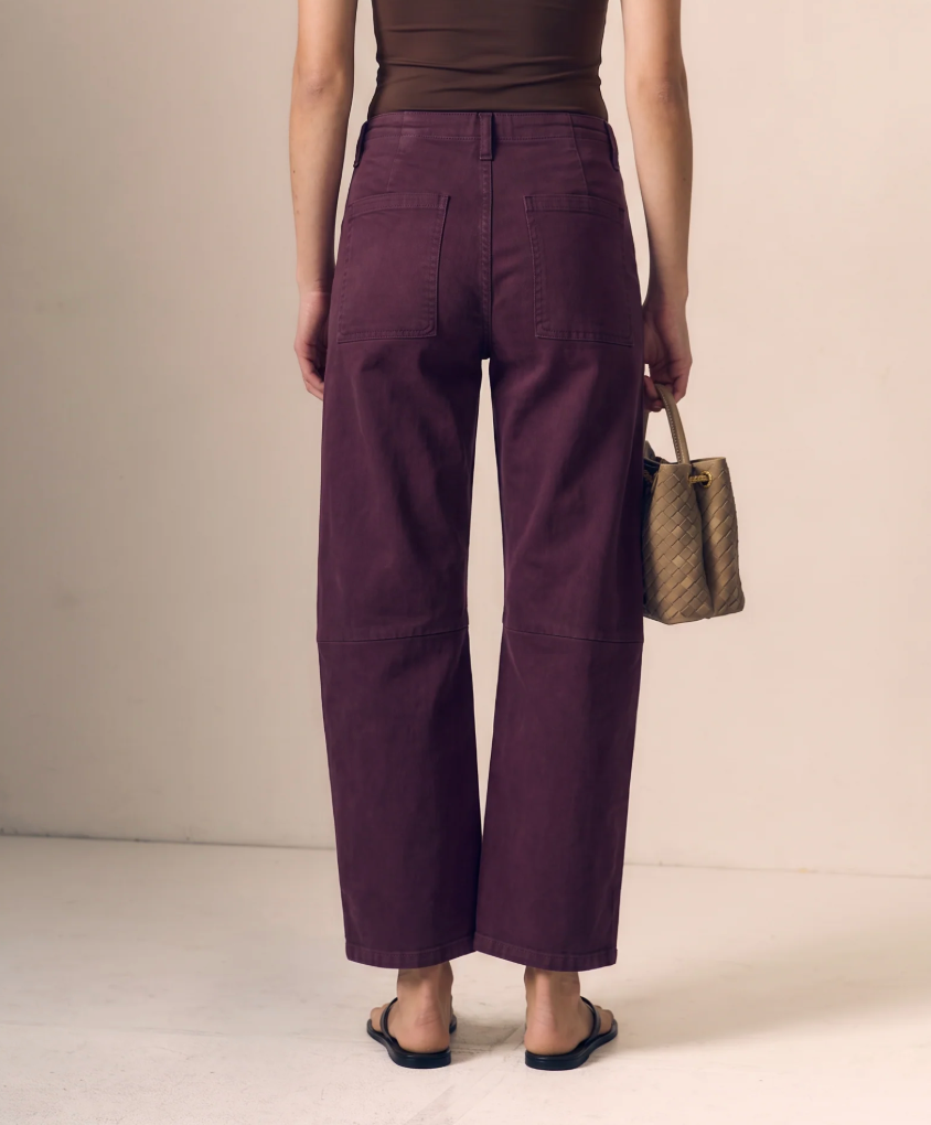 A model shown from the back wearing wide-leg cropped pants in a deep plum purple color, paired with a brown top and black sandals, holding a small beige handbag against a plain beige studio background.