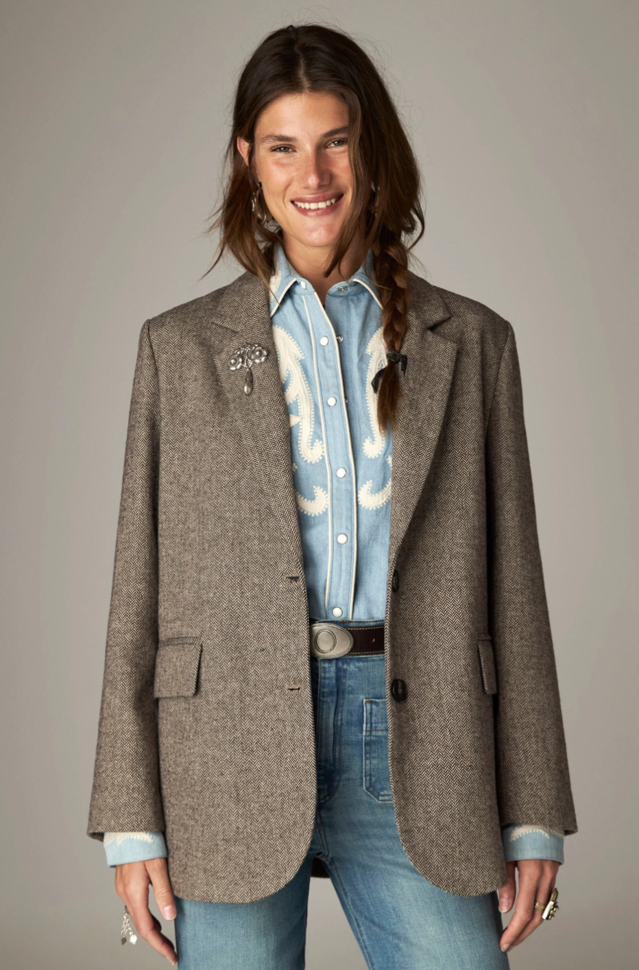 A woman modeling a brown textured tweed blazer over a light blue western embroidered shirt, paired with blue jeans and a silver belt, against a solid gray studio background.