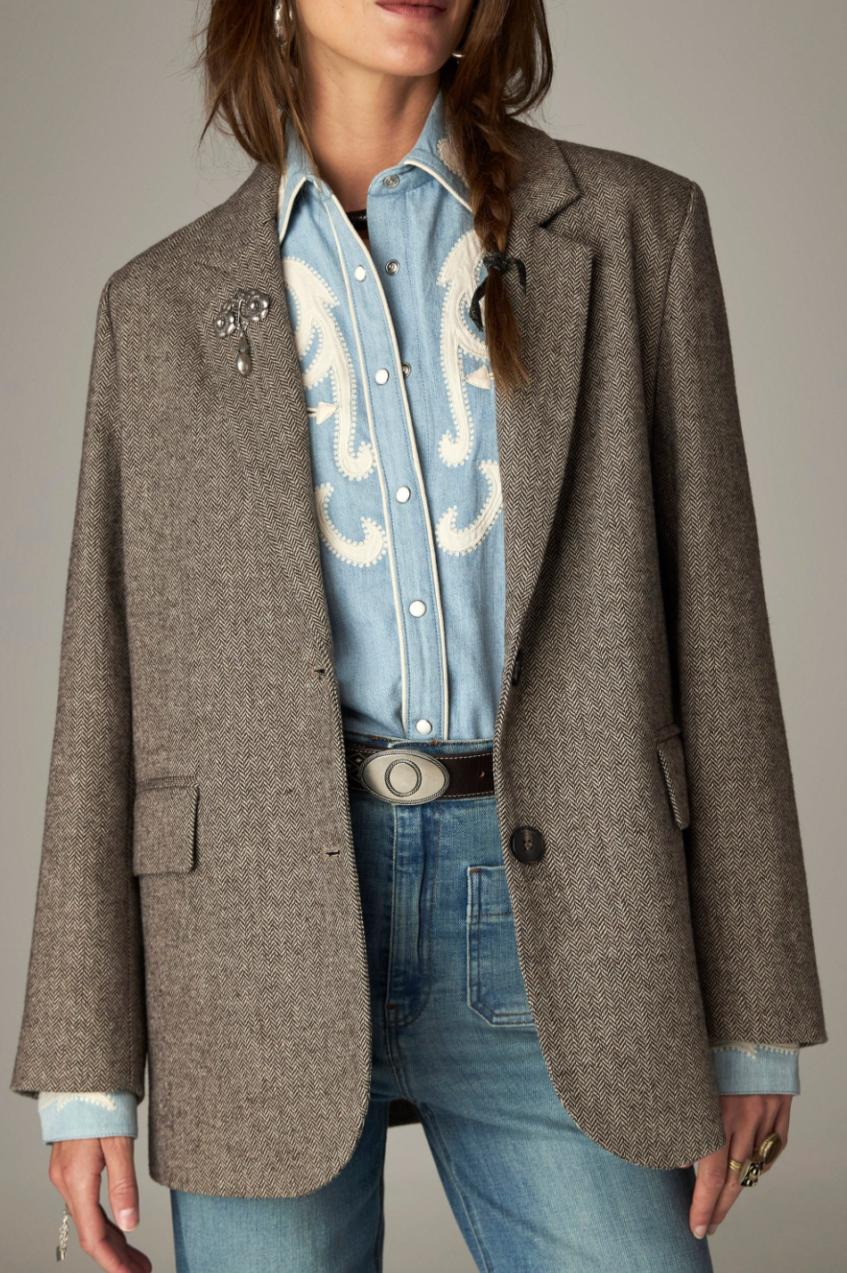 A model wears a brown herringbone tweed blazer over a light blue western-style denim shirt and blue jeans, accessorized with a jeweled brooch and a silver belt.