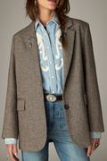 A model wears a brown herringbone tweed blazer over a light blue western-style denim shirt and blue jeans, accessorized with a jeweled brooch and a silver belt.