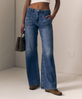 A model wearing a pair of wide-leg, high-waisted blue denim jeans with patch pockets, styled with a white crop top, brown suede shoes, and holding a brown patterned handbag against a minimalist off-white background.