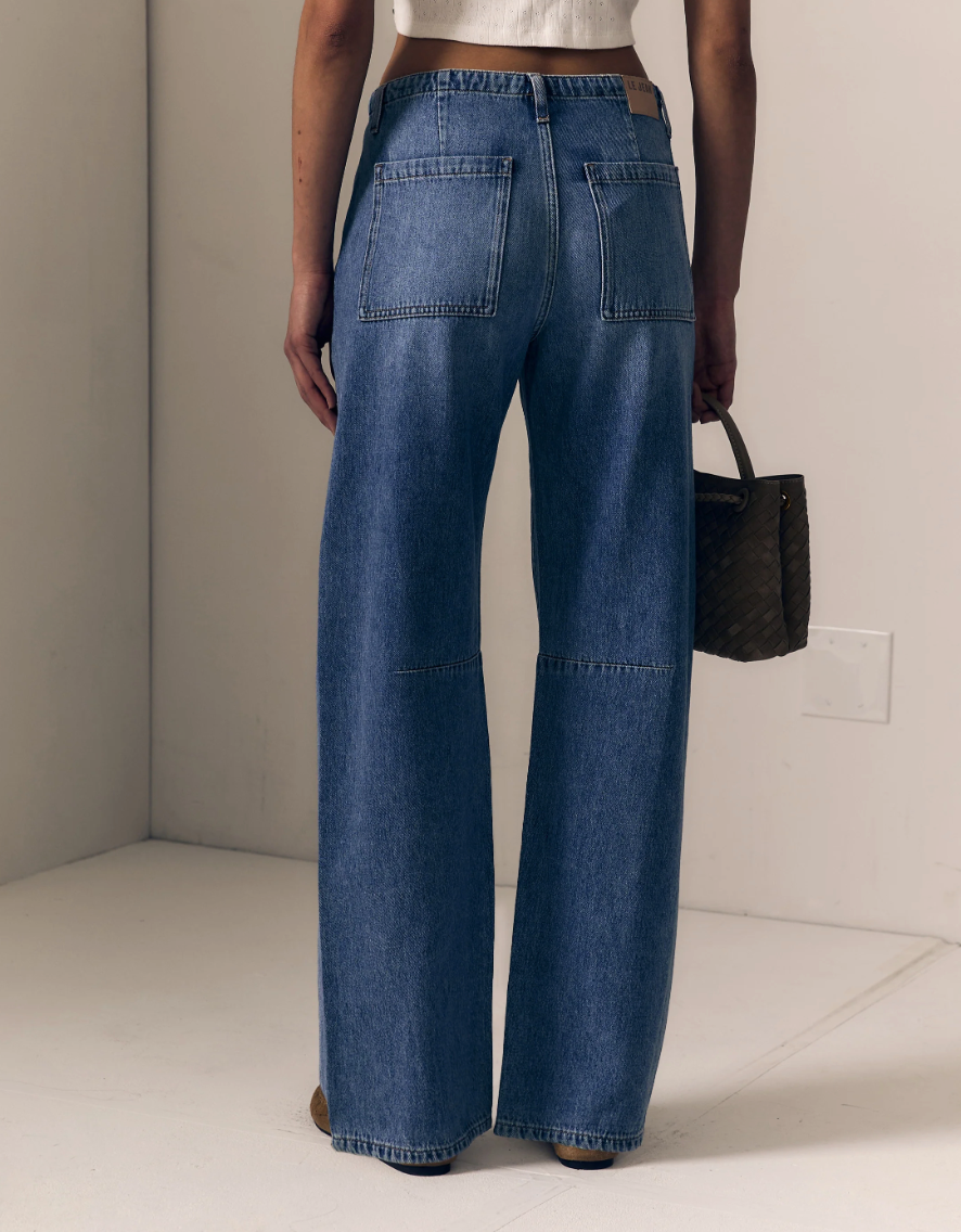 A person shown from the back wearing wide-leg blue denim jeans with patch pockets, standing in a minimalist indoor setting and holding a dark woven handbag.
