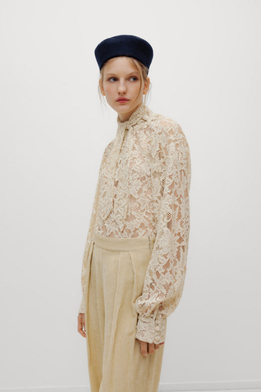 A model wearing a beige lace long-sleeve blouse with a high mock neck, paired with beige trousers and a dark navy blue beret, photographed against a plain white studio background.
