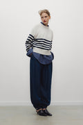 A model stands against a plain white wall wearing a cream mock-neck sweater with navy horizontal stripes, a blue plaid shirt underneath, loose dark blue trousers, and black pointed shoes.