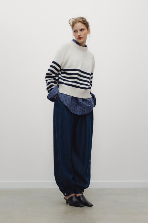 A model stands against a plain white wall wearing a cream mock-neck sweater with navy horizontal stripes, a blue plaid shirt underneath, loose dark blue trousers, and black pointed shoes.