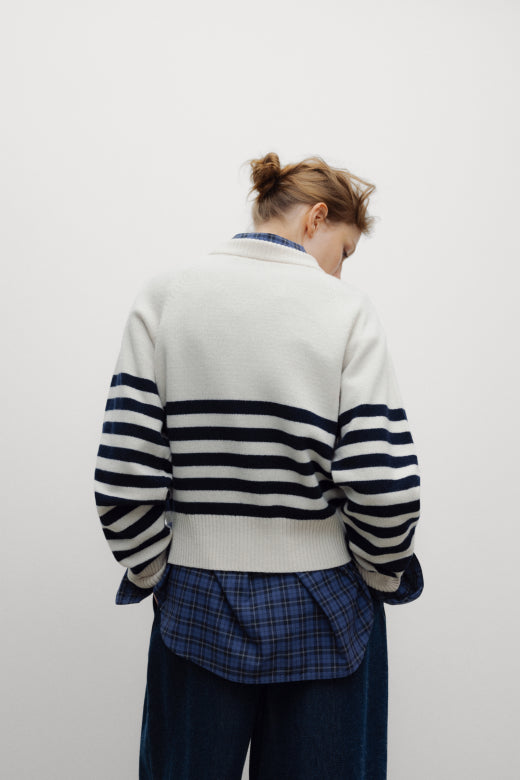 A person viewed from behind wearing a cream knit cardigan with navy horizontal stripes across the upper back and sleeves, layered over a blue plaid shirt and dark blue pants, against a plain light gray background.
