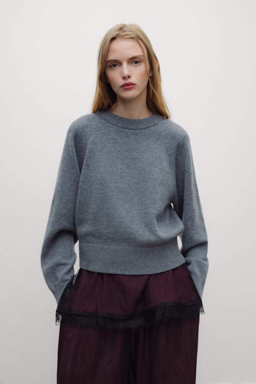 A model wearing a heather gray crewneck sweater and a dark burgundy skirt with black lace trim, standing against a plain light gray background.
