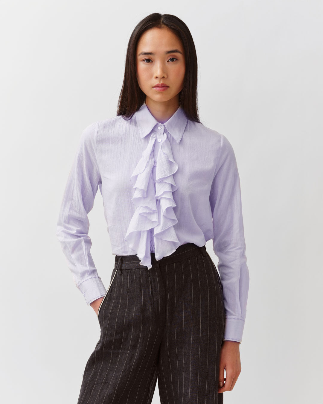 Woman wearing a light purple blouse with ruffles and dark pinstripe pants on a white background